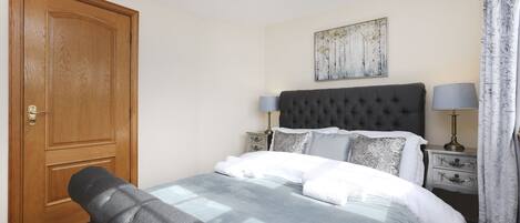 A neatly arranged bedroom with a large bed, a wooden door, a bedside table with lamps, and a wall-mounted artwork.