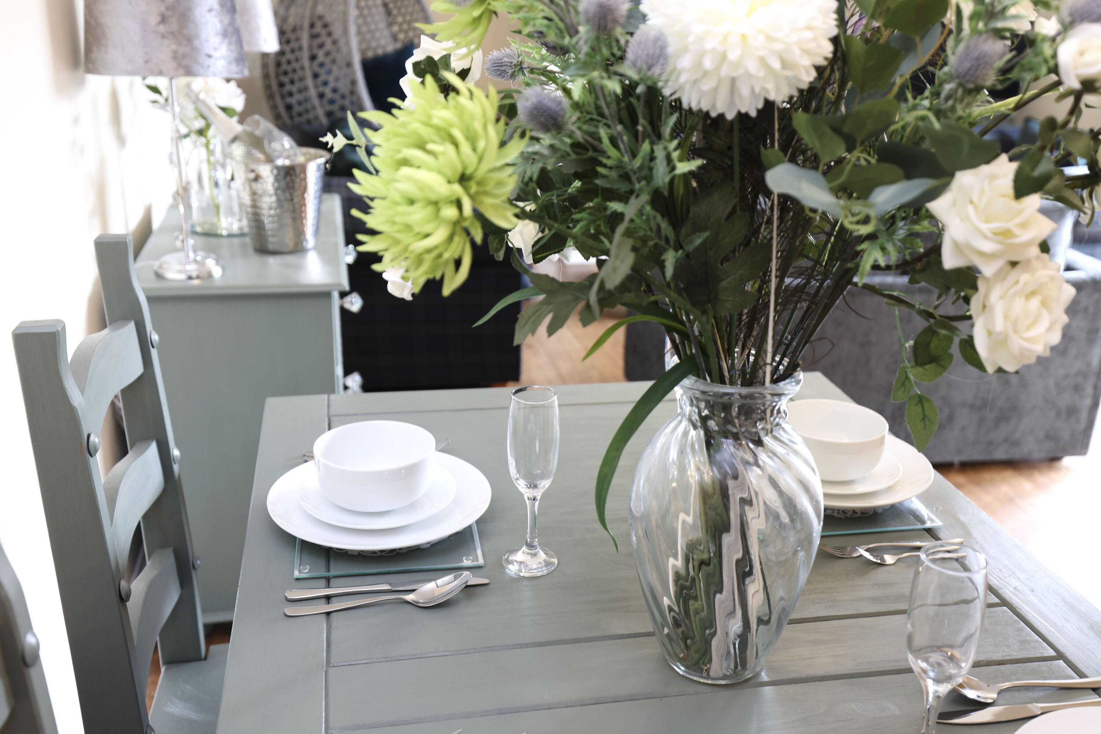 A dining table set with white plates, silverware, and a clear vase with flowers.