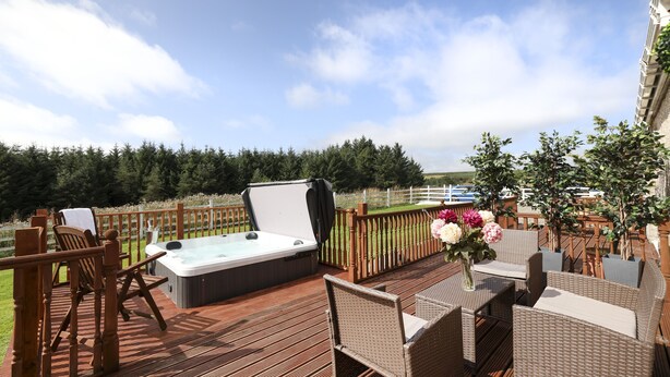 A wooden deck with a hot tub, wicker furniture, and a view of a lake. - JOIVY Greenknowes Lodge With Hot Tub And Wood Burning Stove (Kelty)