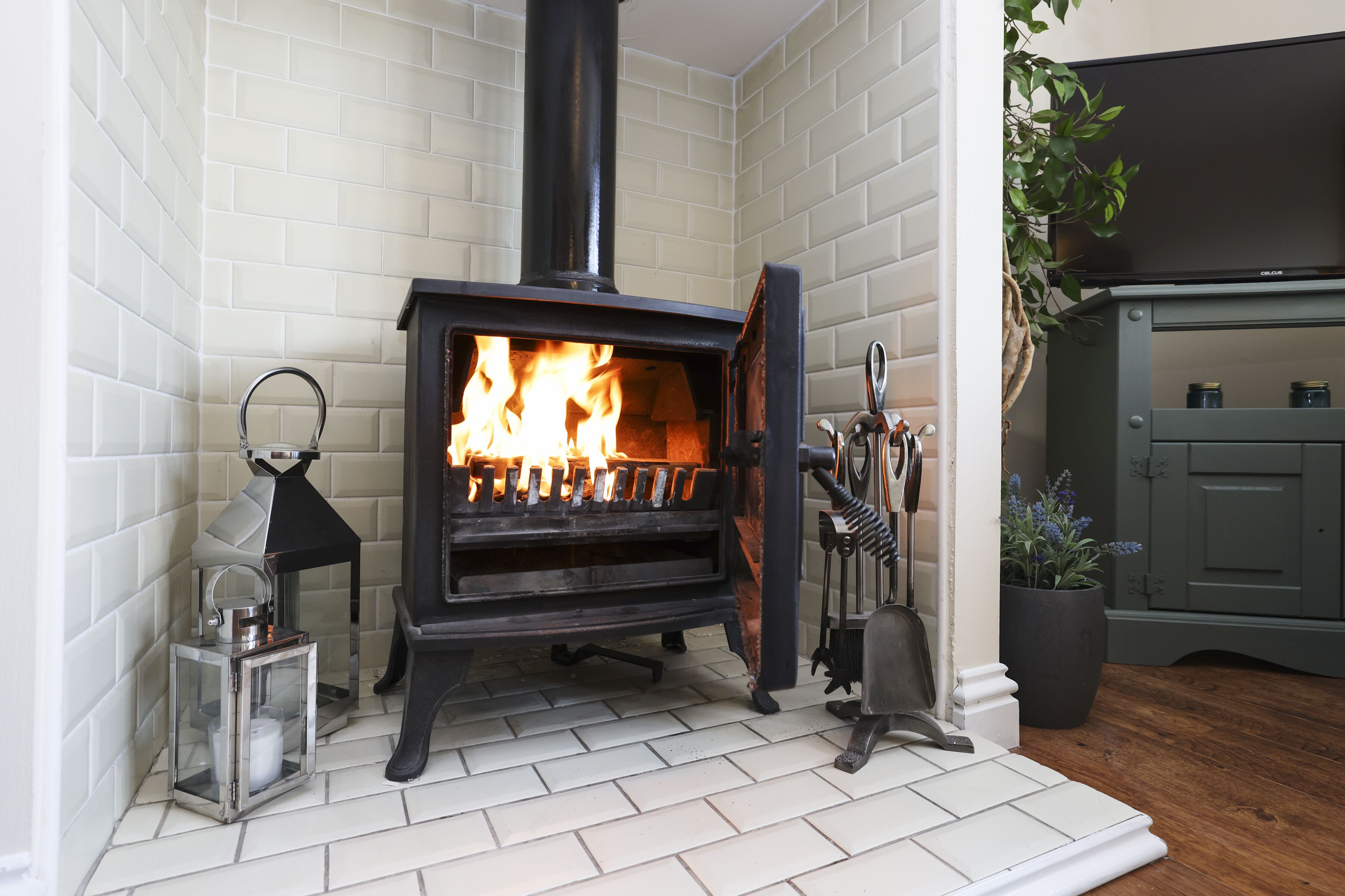 Un poêle à bois avec un feu, une lanterne et une plante en pot, le tout placé dans un coin d’une pièce aux murs recouverts de carreaux blancs et au plancher en bois.