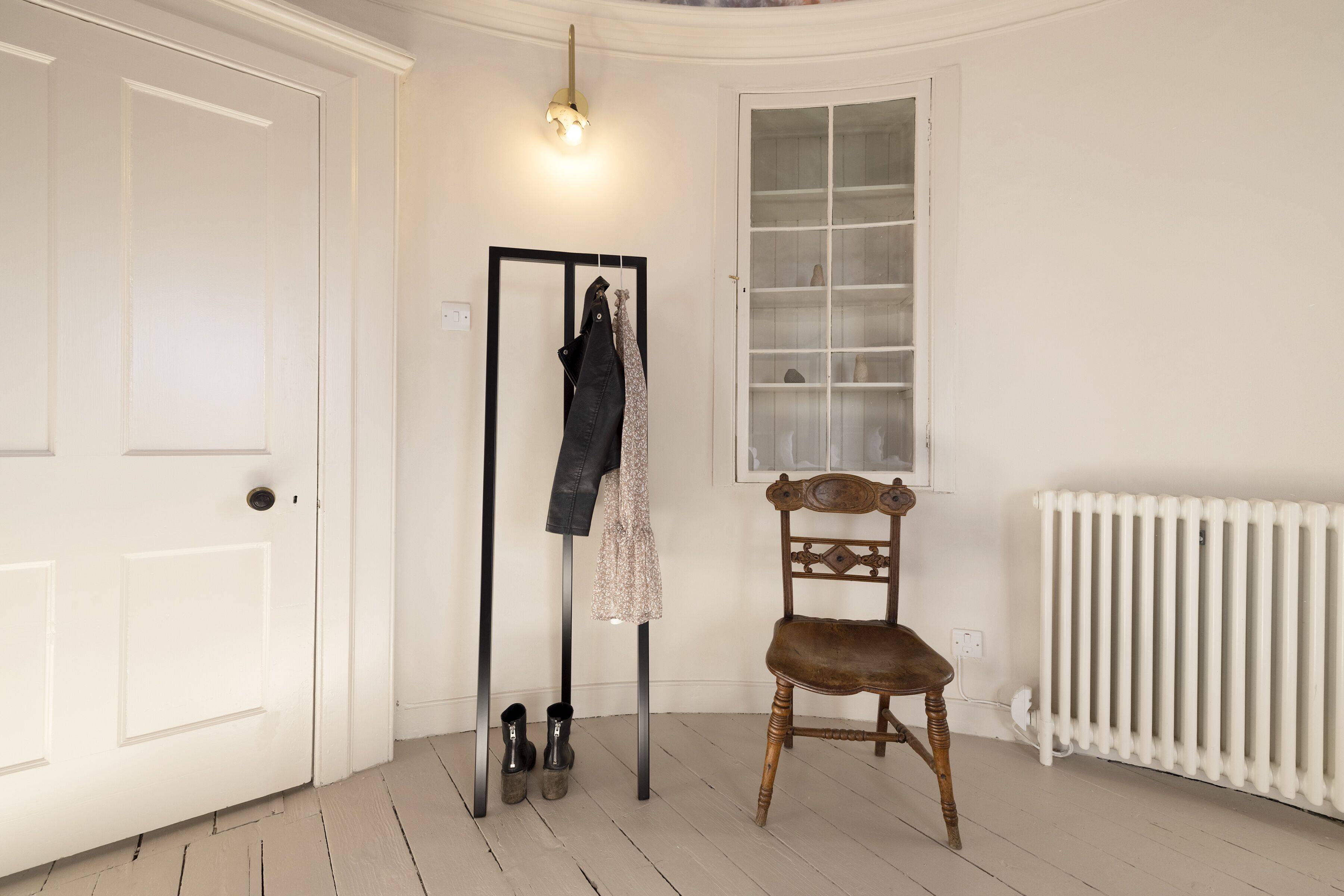 A room with a white door, a coat rack with clothes, a wooden chair, and a radiator.