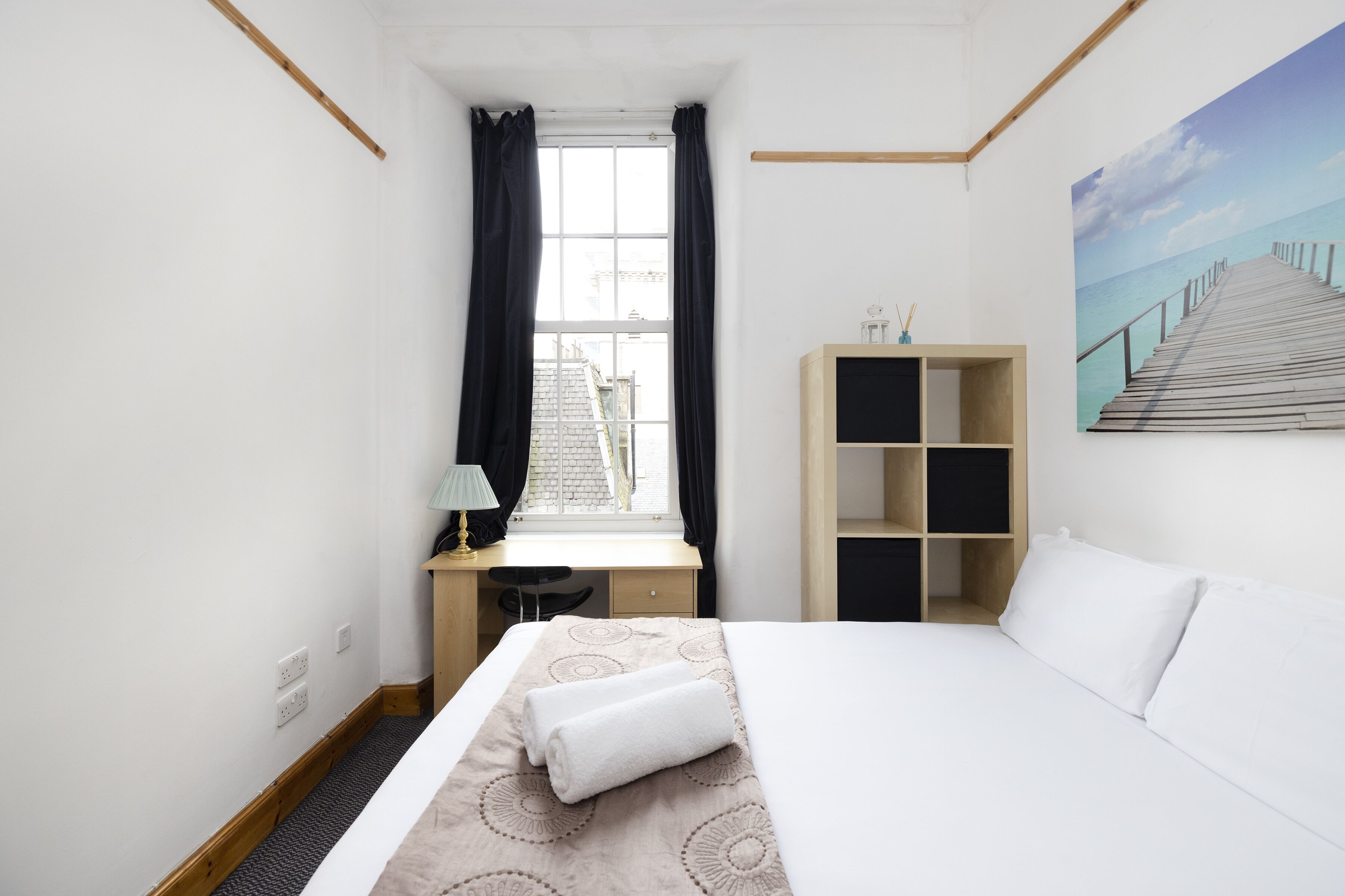 A hotel room with a bed, a desk, a chair, a window with curtains, a bookshelf, and a painting of a pier.