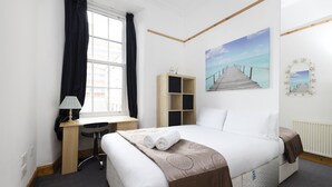 A hotel room with a bed, desk, chair, and a painting of a pier. - JOIVY Spacious 3 Bed Apartment In The Old Town (Edinburgh)