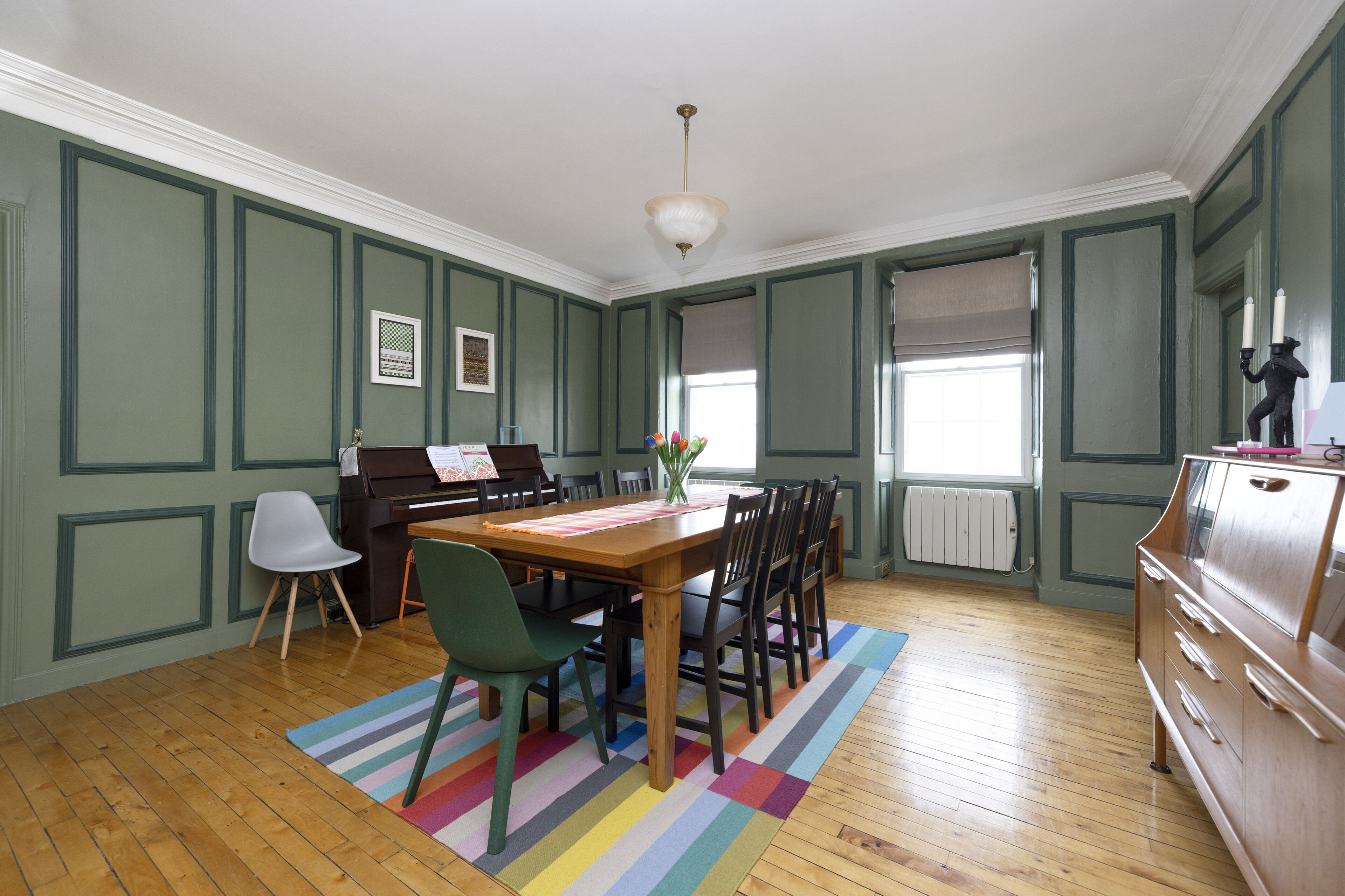 Uma sala de jantar com um piano, uma mesa com cadeiras, um tapete colorido e um aparador.