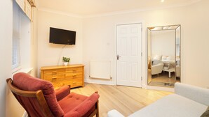 Une chambre avec une commode en bois, un téléviseur fixé au mur, un grand miroir et une porte donnant sur une autre pièce.