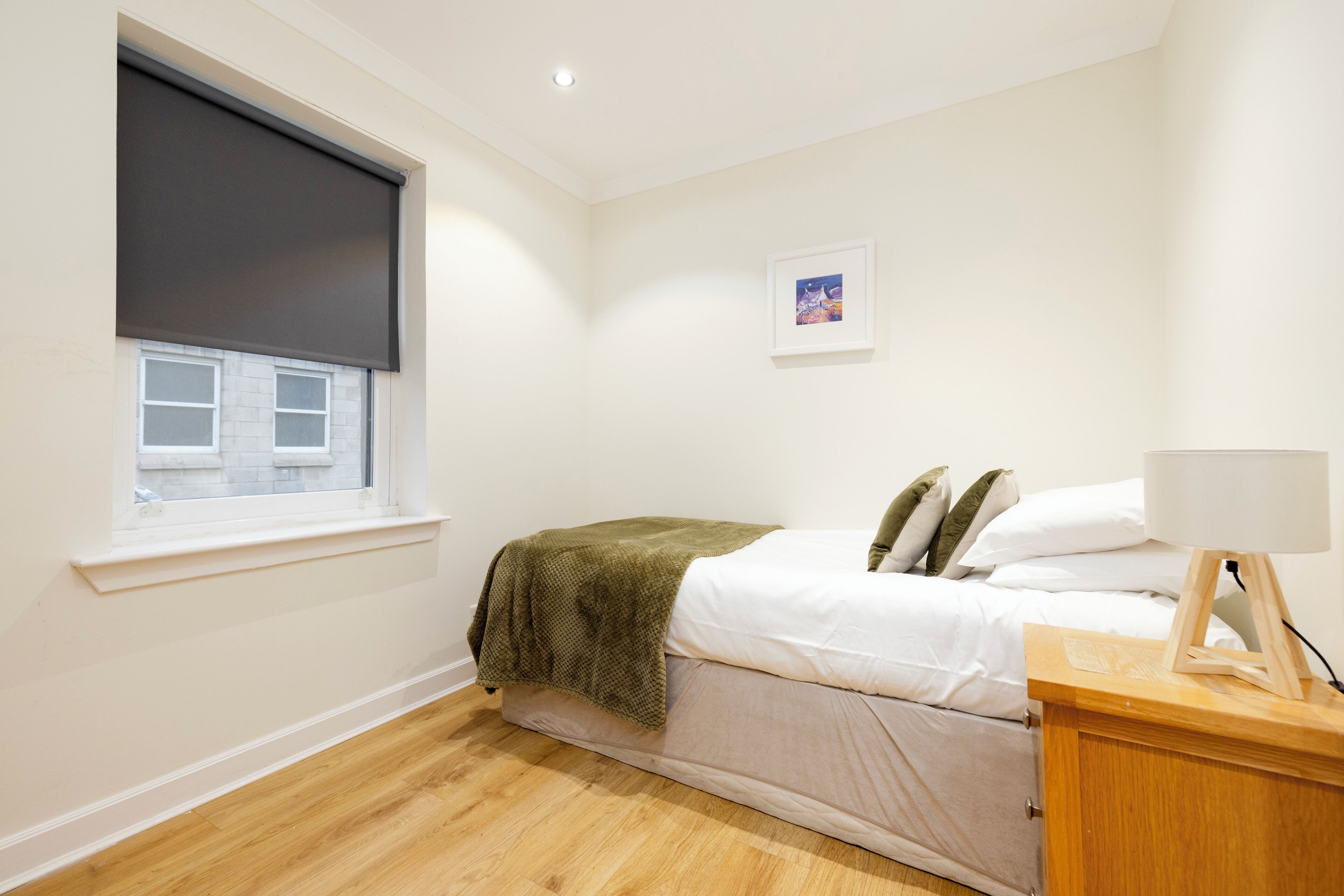A bedroom with a bed, a bedside table, a window with a blind, a picture on the wall, and a wooden floor.