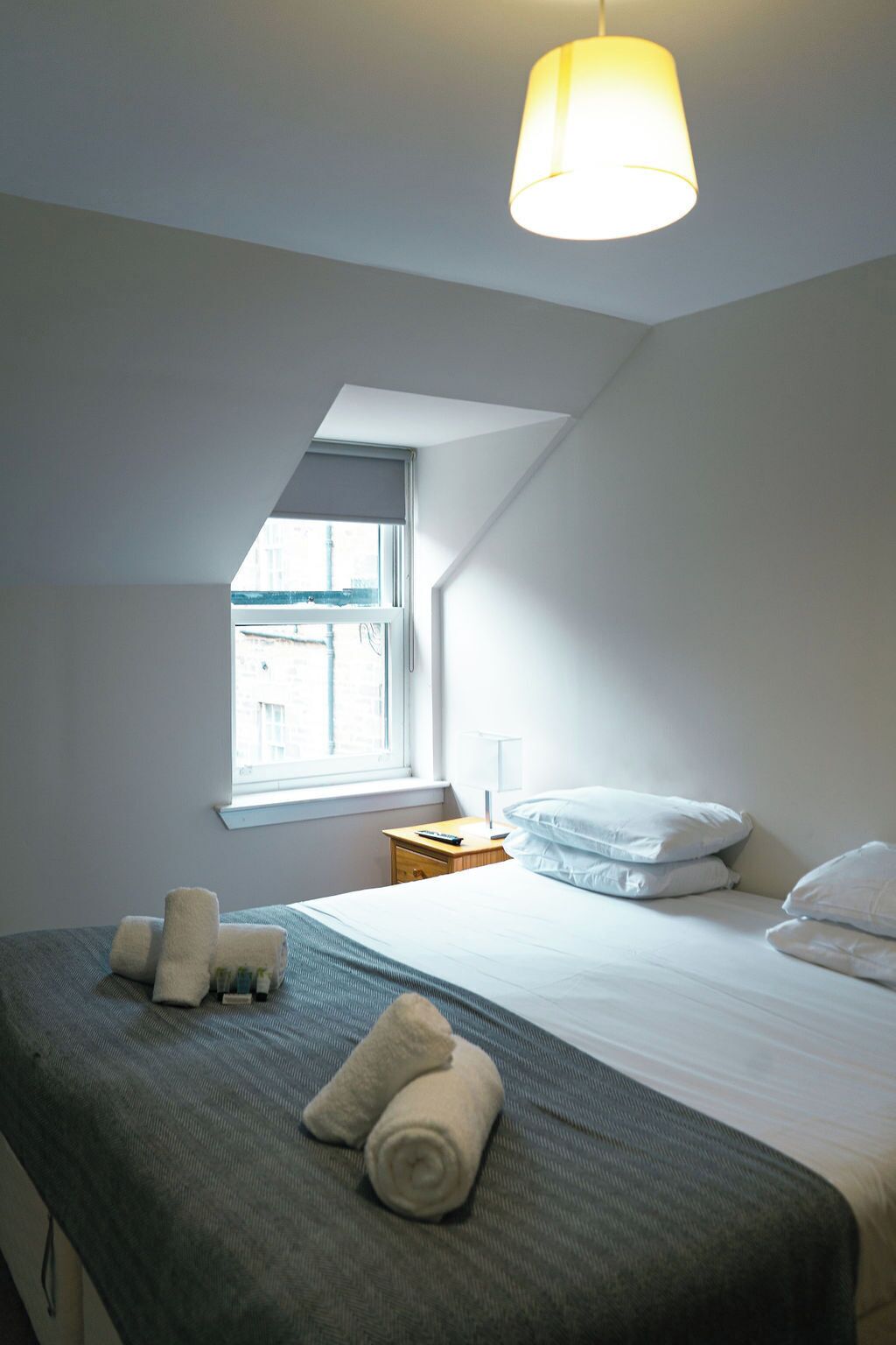Un dormitorio con cama, almohadas, una mesita de noche, una lámpara y una ventana.