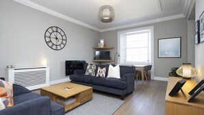 A modern living room with a sofa, coffee table, and a TV. There is a window providing natural light and a decorative lamp. - JOIVY Splendid 2-Br Apartment In New Town (Edinburgh)