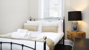 A neatly made bed with white linens and a wooden headboard, a bedside table with a lamp, and a window with blinds. - JOIVY Modern 1-Br Apt W/ Communal Terrace In Trendy Stockbridge (Edinburgh)
