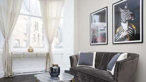 A modern living room with a grey velvet sofa, a glass coffee table, and two framed artworks on the wall. - JOIVY Modern 1-Br Apt W/ Communal Terrace In Trendy Stockbridge (Edinburgh)