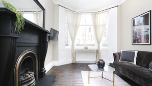 A living room with a fireplace, a dark sofa, a coffee table, and a large window with curtains. - JOIVY Modern 1-Br Apt W/ Communal Terrace In Trendy Stockbridge (Edinburgh)