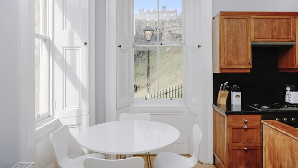 A kitchen with a white table and chairs, wooden cabinets, and a view of a castle through a window.
