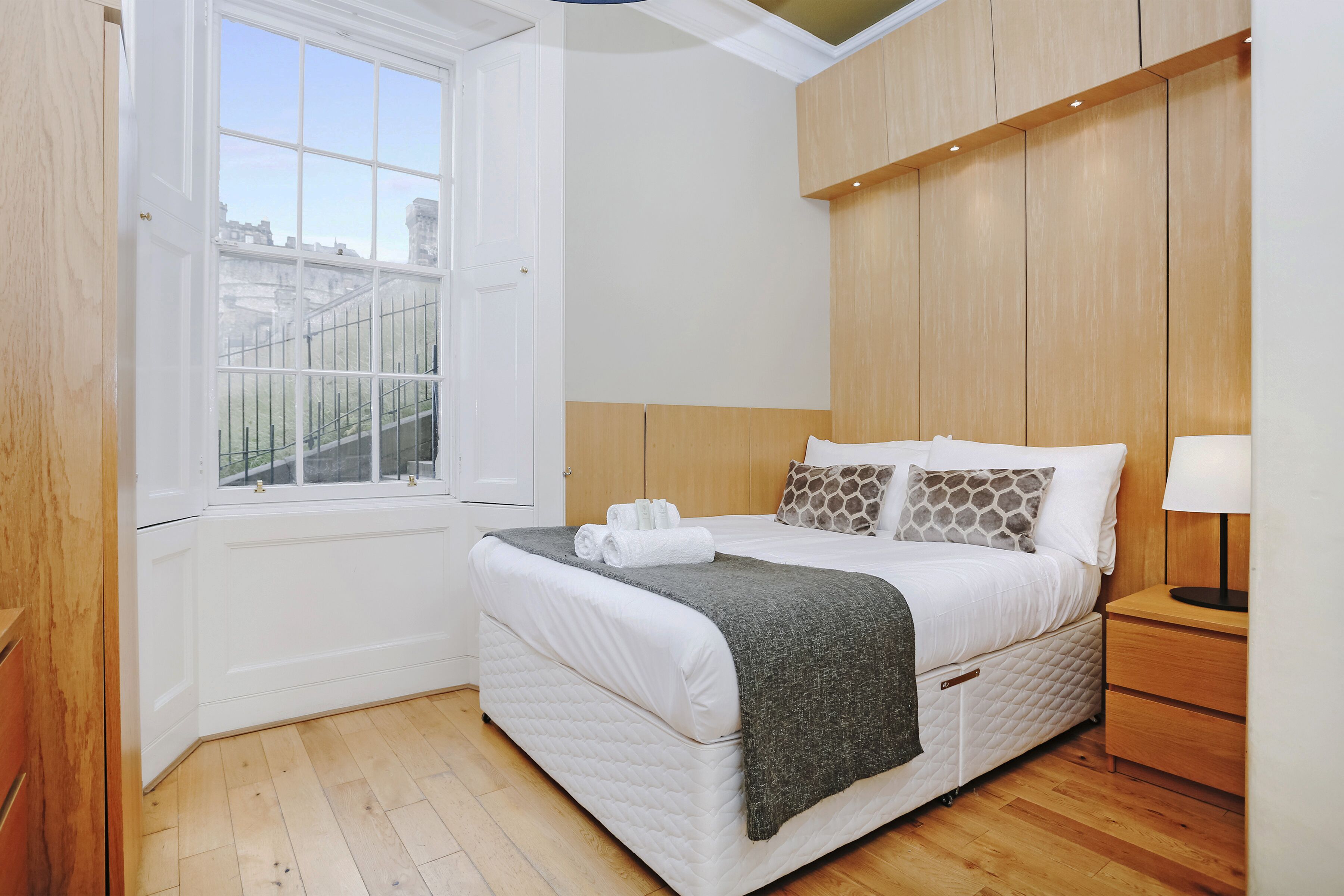 Ein Schlafzimmer mit großem Fenster, einem Bett mit weißer Bettwäsche und einer gesteppten Matratze, einem hölzernen Nachttisch und einer Lampe.