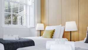 A hotel room with two beds, a wooden headboard, bedside lamps, and a window with a view of a building and a staircase.