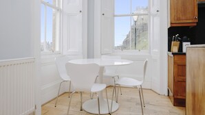 A bright kitchen with white cabinetry, a round dining table with four chairs, and large windows offering a view of a cityscape.