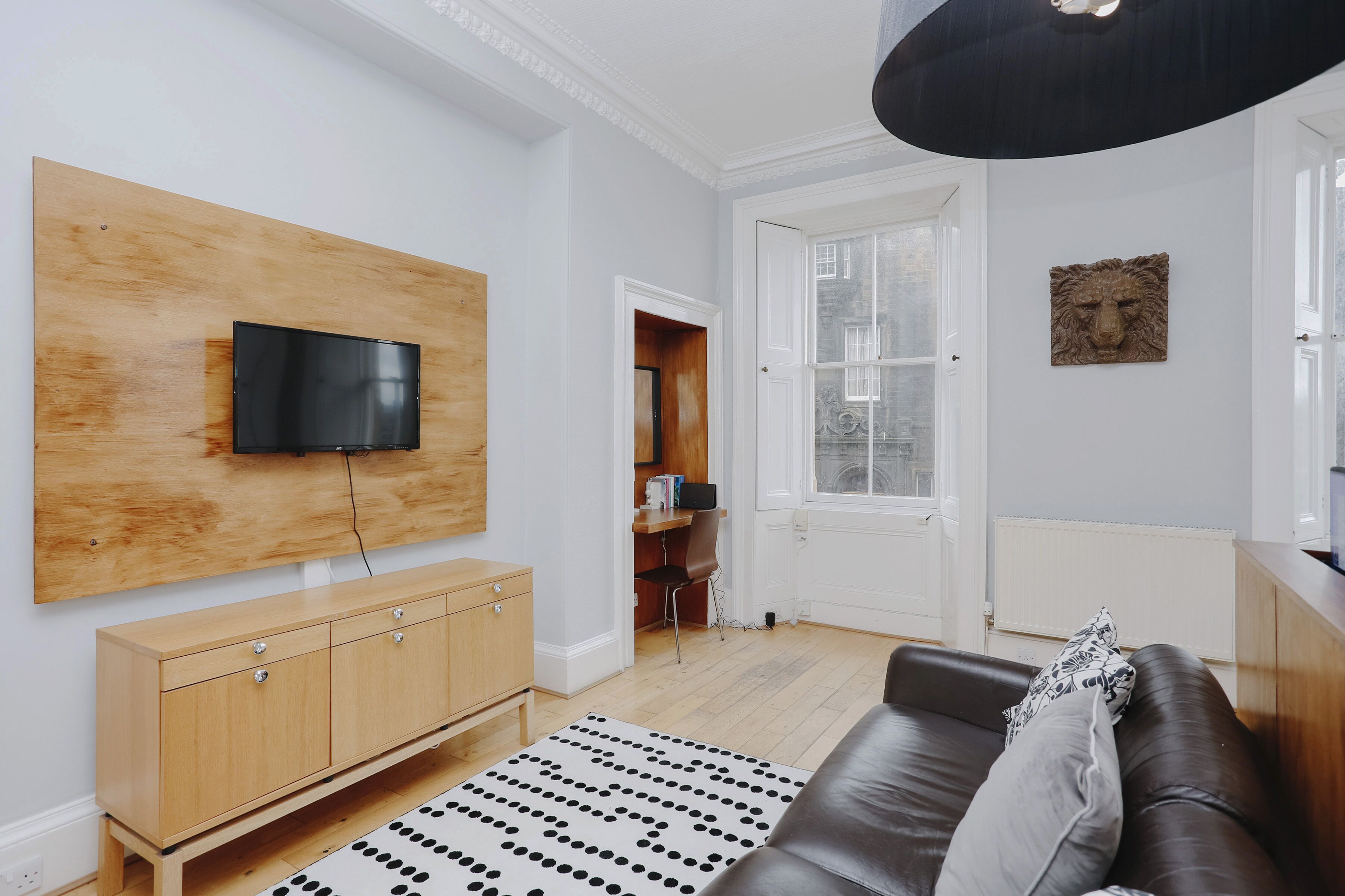 A modern living room with a flat-screen TV mounted on a wooden panel, a leather sofa, a patterned rug, and a wooden cabinet.