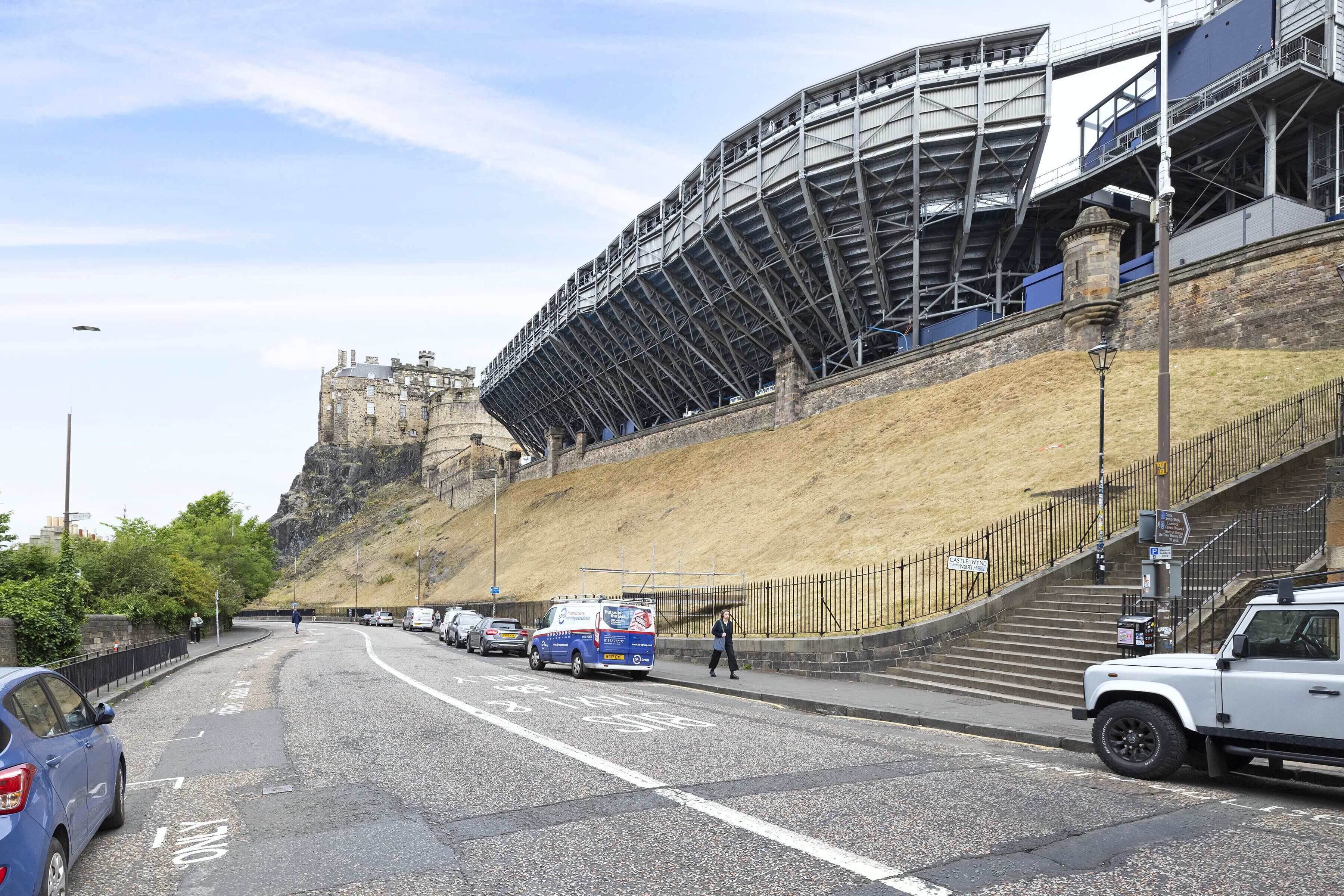 A stadium with a steep hillside, a parking area, and a pedestrian walkway.
