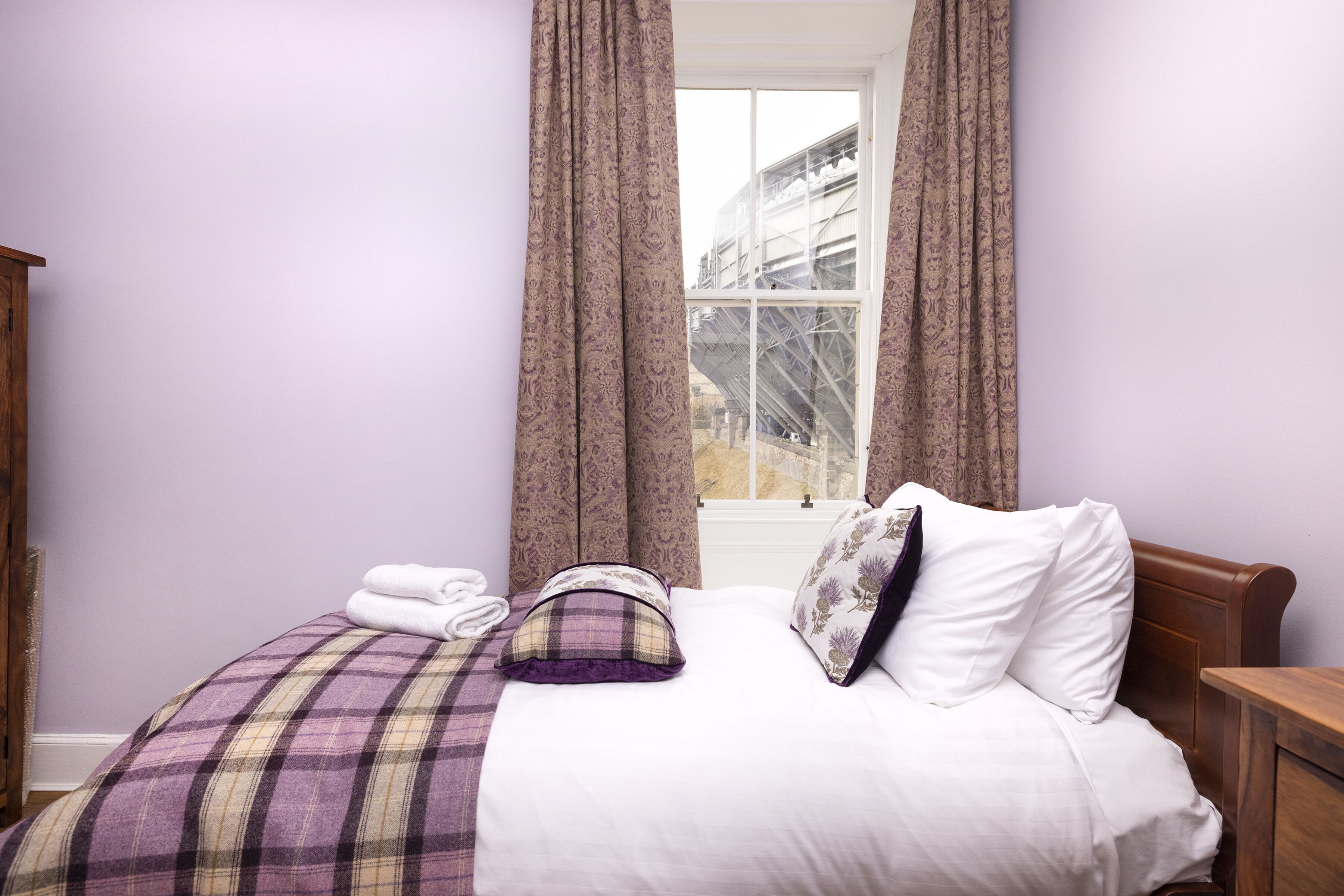 A neatly made bed with a plaid bedspread and white pillows, a window with curtains, and a wooden nightstand.