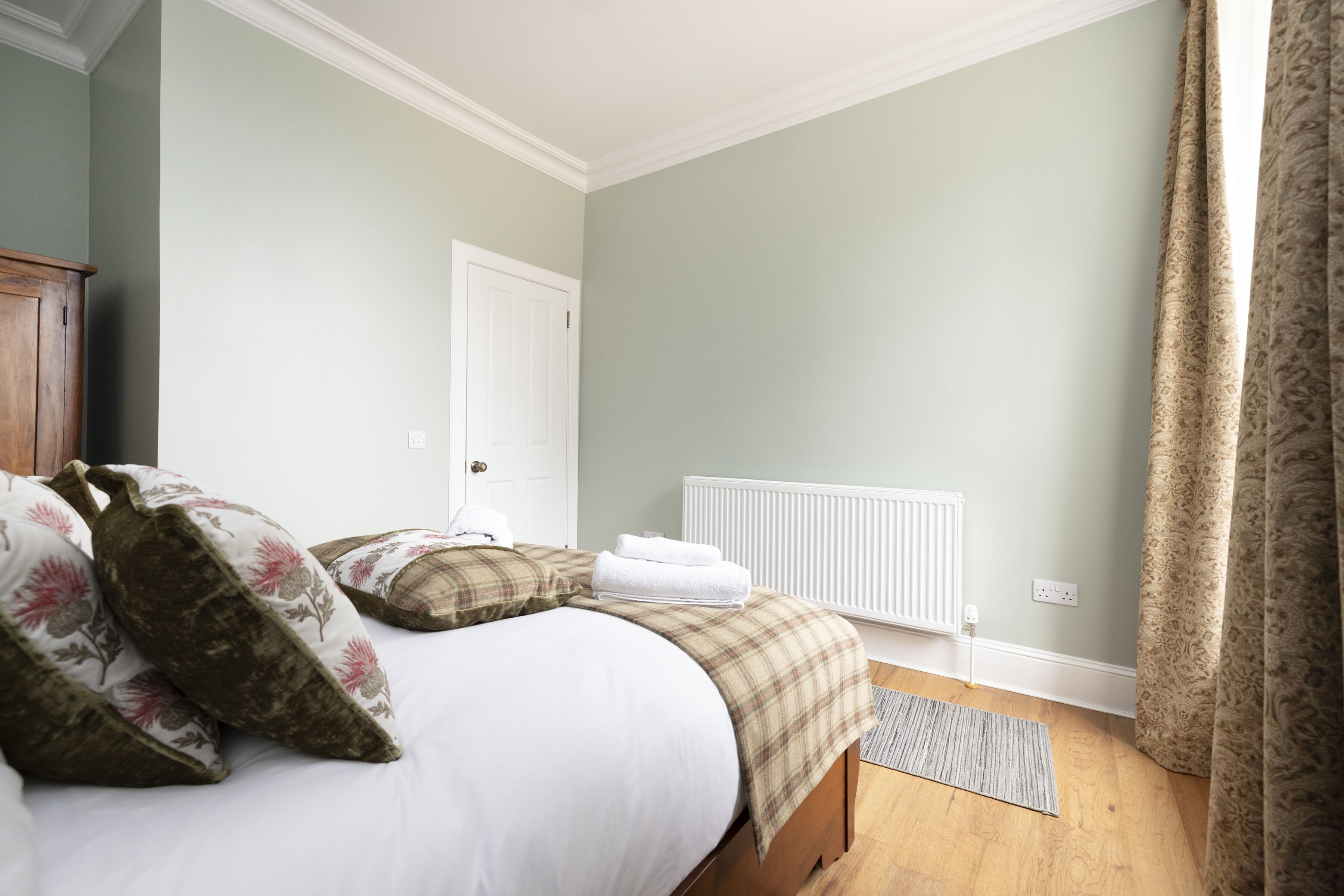 Une chambre à coucher avec un lit, des oreillers, un radiateur, une porte et une fenêtre avec des rideaux.