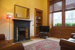 A living room with a fireplace, a television on a stand, a bookshelf, and a large window with blinds.