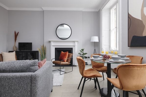 A modern living room with a fireplace, a dining area with orange chairs, and a grey sofa.