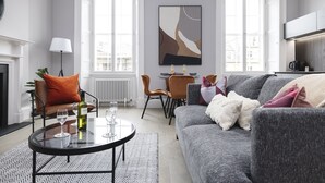 Una sala de estar moderna con un sofá gris, una mesa de centro redonda y una chimenea. Hay dos ventanas que dejan entrar luz natural, y un comedor con mesa y sillas.