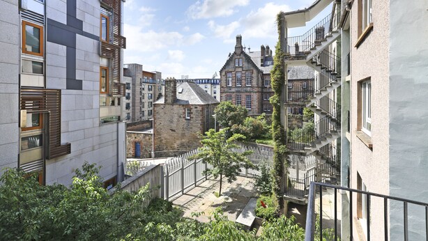 Une vue d’une rue étroite en milieu urbain, bordée de bâtiments des deux côtés, dont un bâtiment en pierre avec un toit en pente et un bâtiment moderne doté de grandes fenêtres et de balcons.