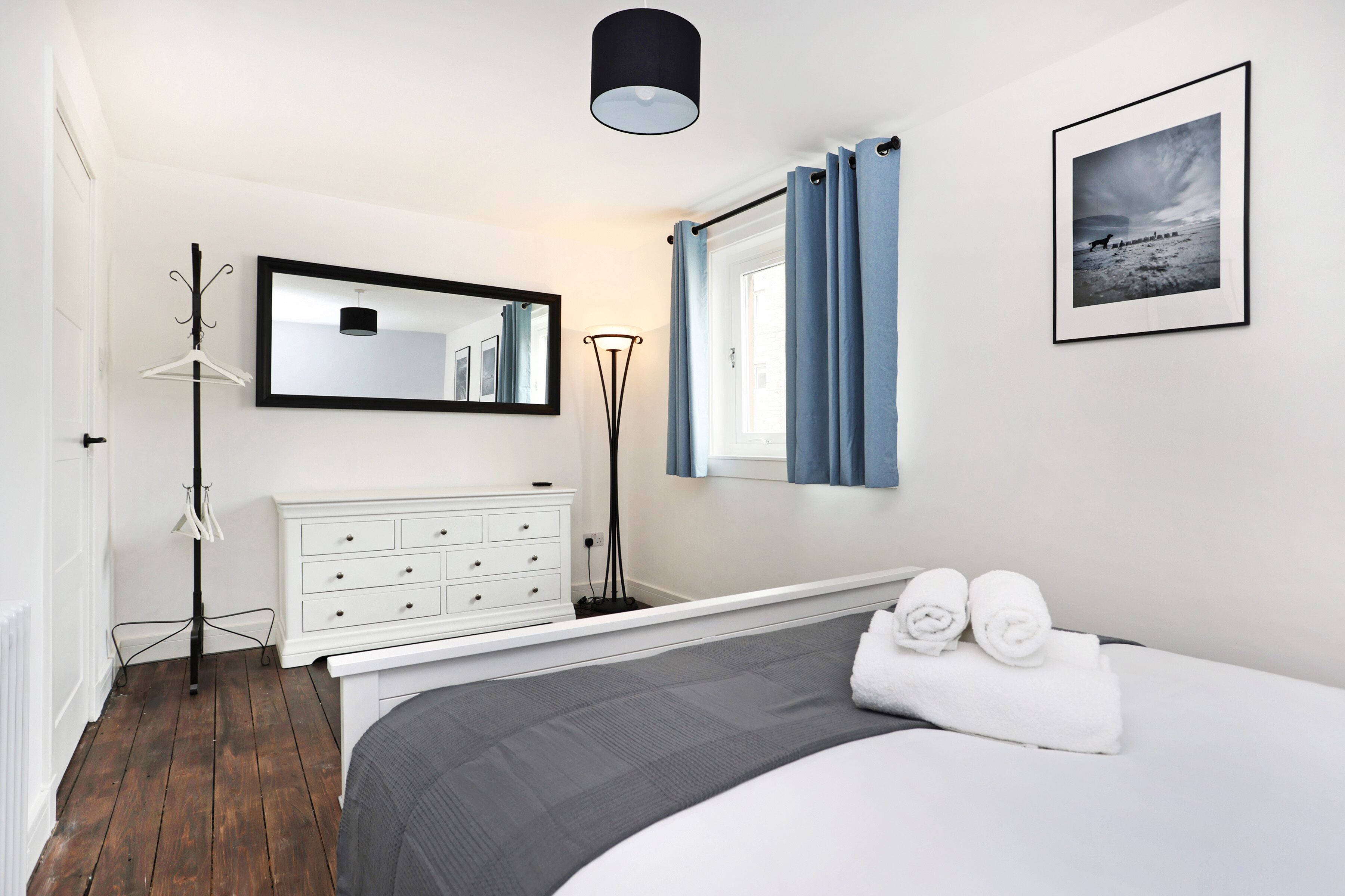 A bedroom with a bed, a dresser, a mirror, a floor lamp, and a framed picture on the wall.