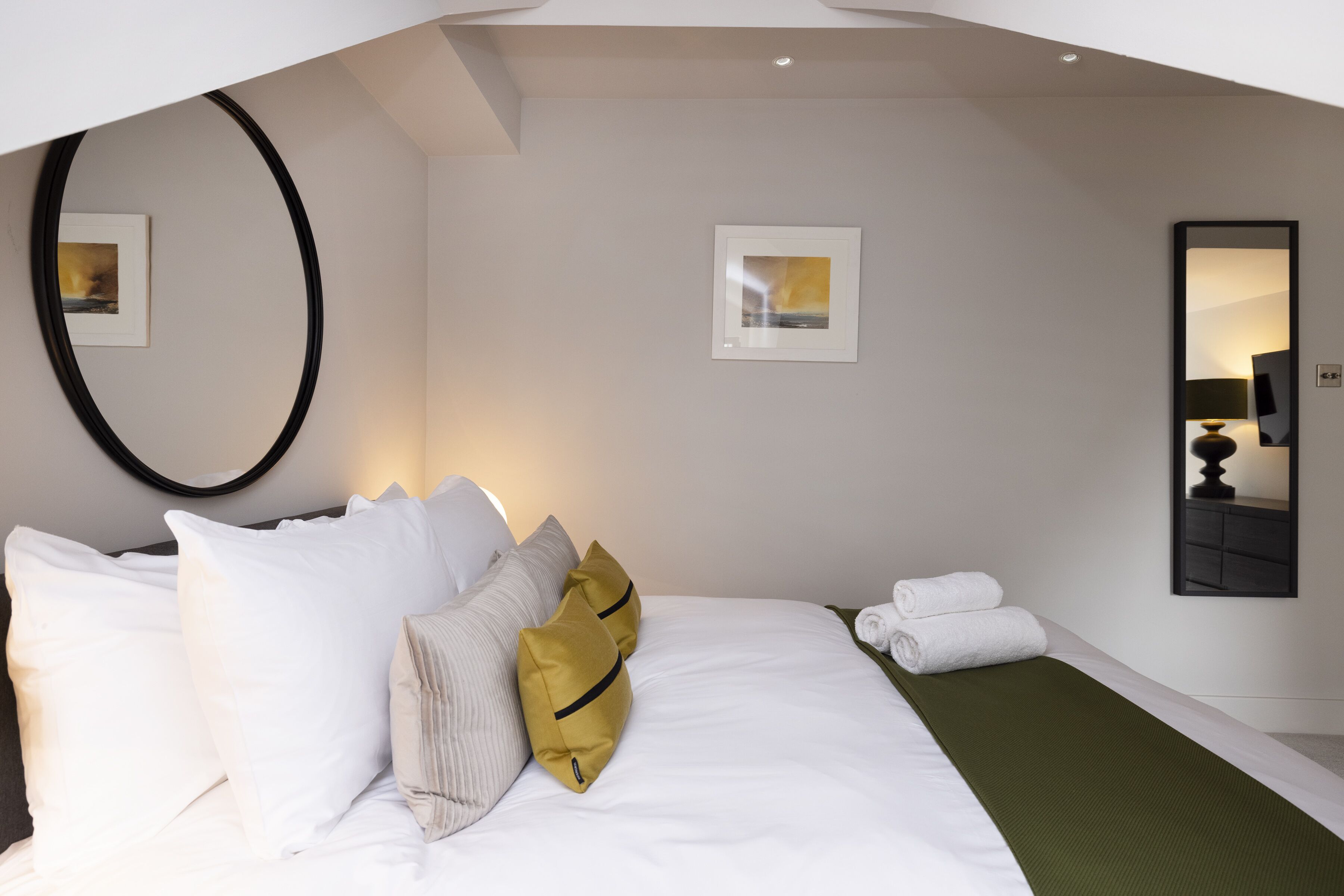 A neatly made bed with white and mustard pillows, a green throw blanket, and a small bedside table with a lamp.