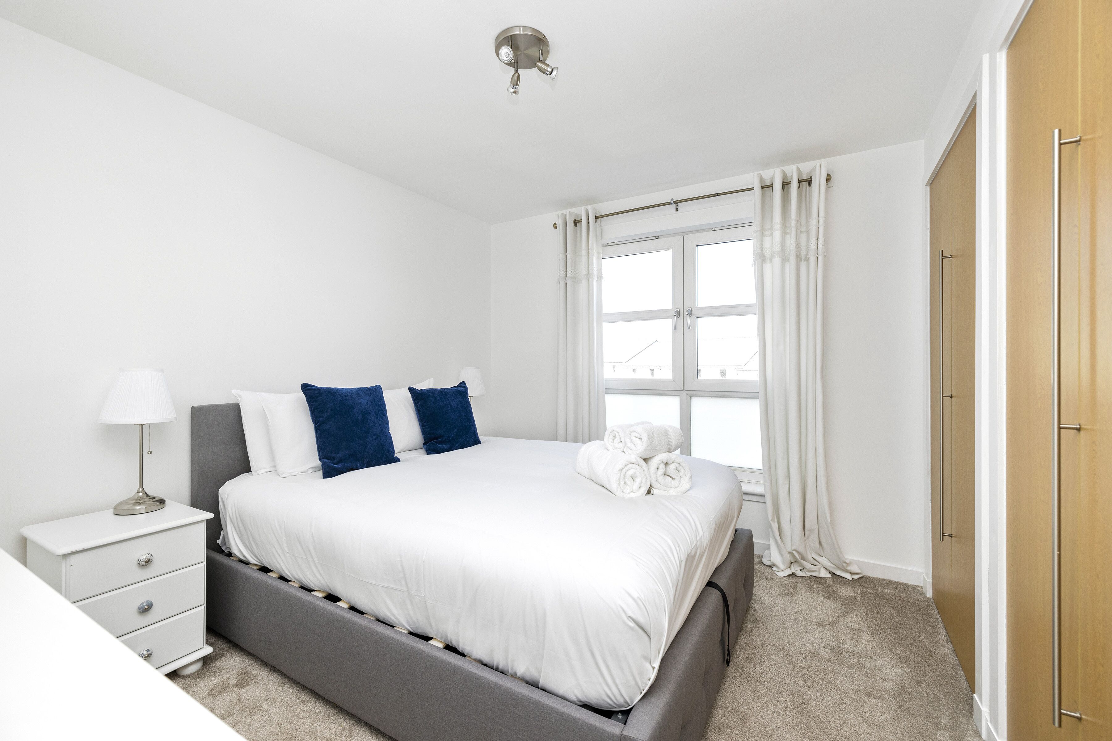A bedroom with a bed, a nightstand, a lamp, a window with curtains, and a wardrobe.