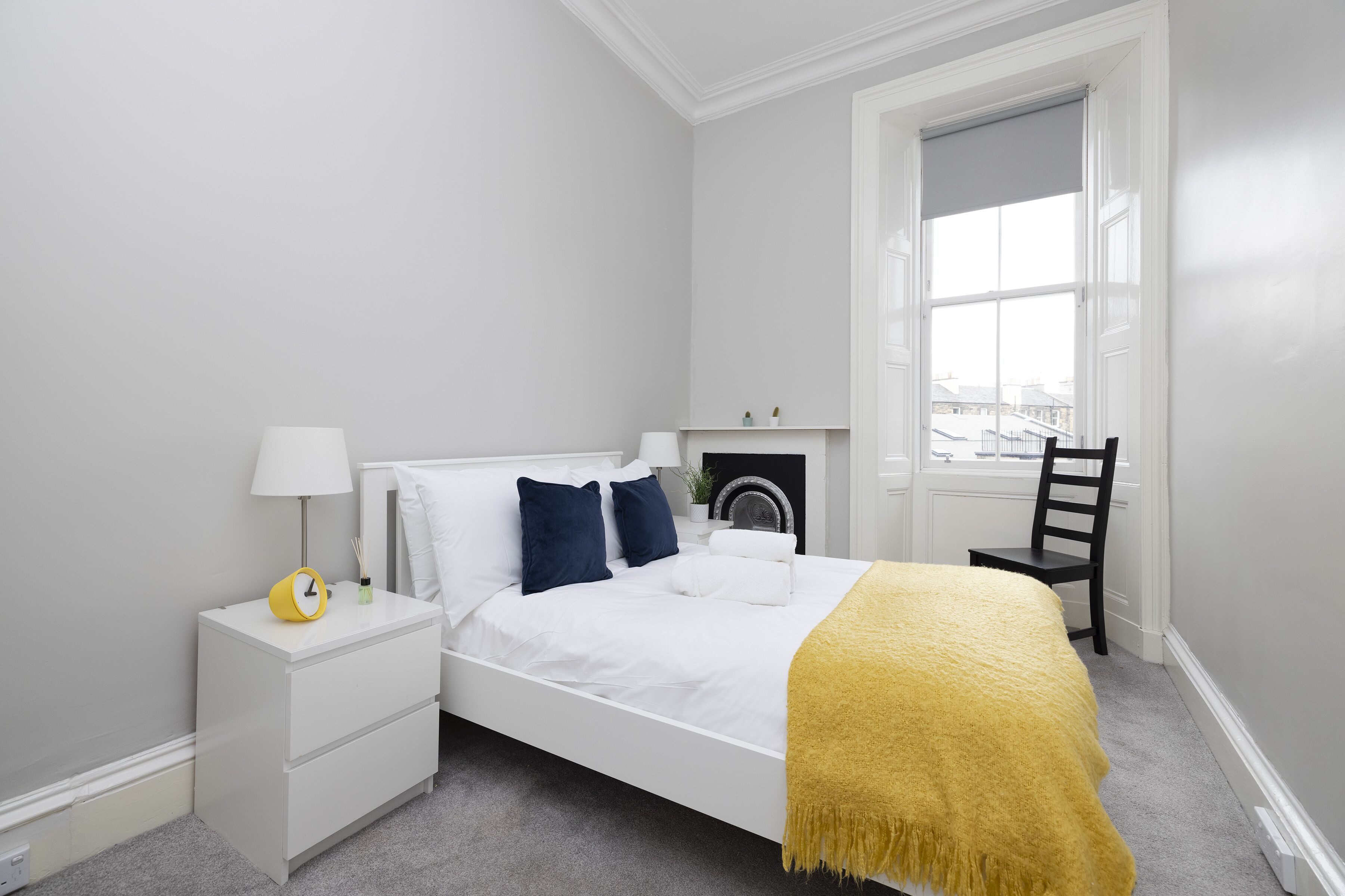 Un dormitorio ordenado con cama, mesita de noche, lámpara y una silla.