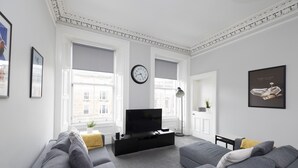 A modern living room with a large grey sofa, a black entertainment unit, and a flat-screen TV. There are large windows with blinds, a clock on the wall, and a framed picture on the wall. - JOIVY 4-Bed Stylish Flat In Edinburgh City Centre (Edinburgh)