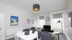 A modern living room with a grey sofa, white dining table, and a flat-screen TV. - JOIVY 4-Bed Stylish Flat In Edinburgh City Centre (Edinburgh)