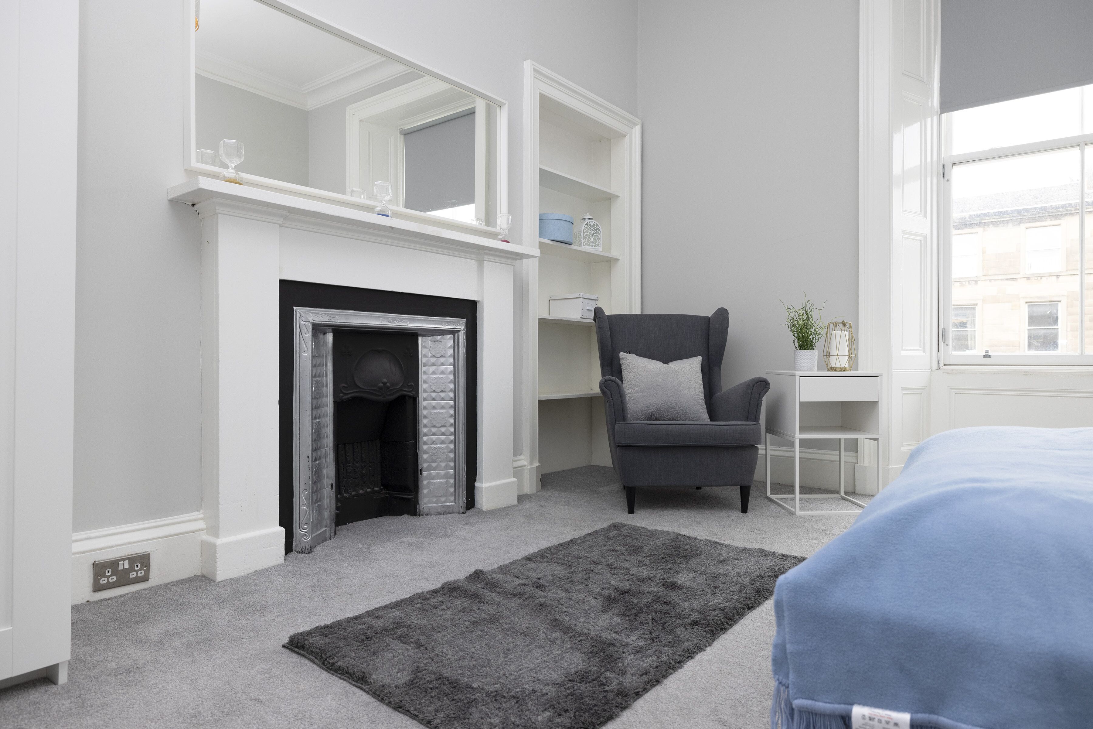 A modern bedroom with a fireplace, a grey armchair, a small table, and a large window.