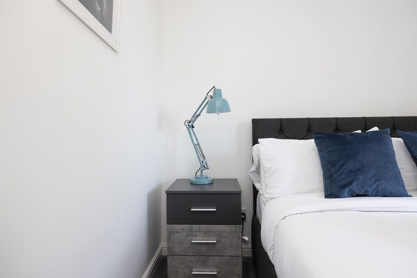 Una cama bien hecha con almohadas blancas y azules, una mesita de noche gris con cajones, y una lámpara de escritorio azul.