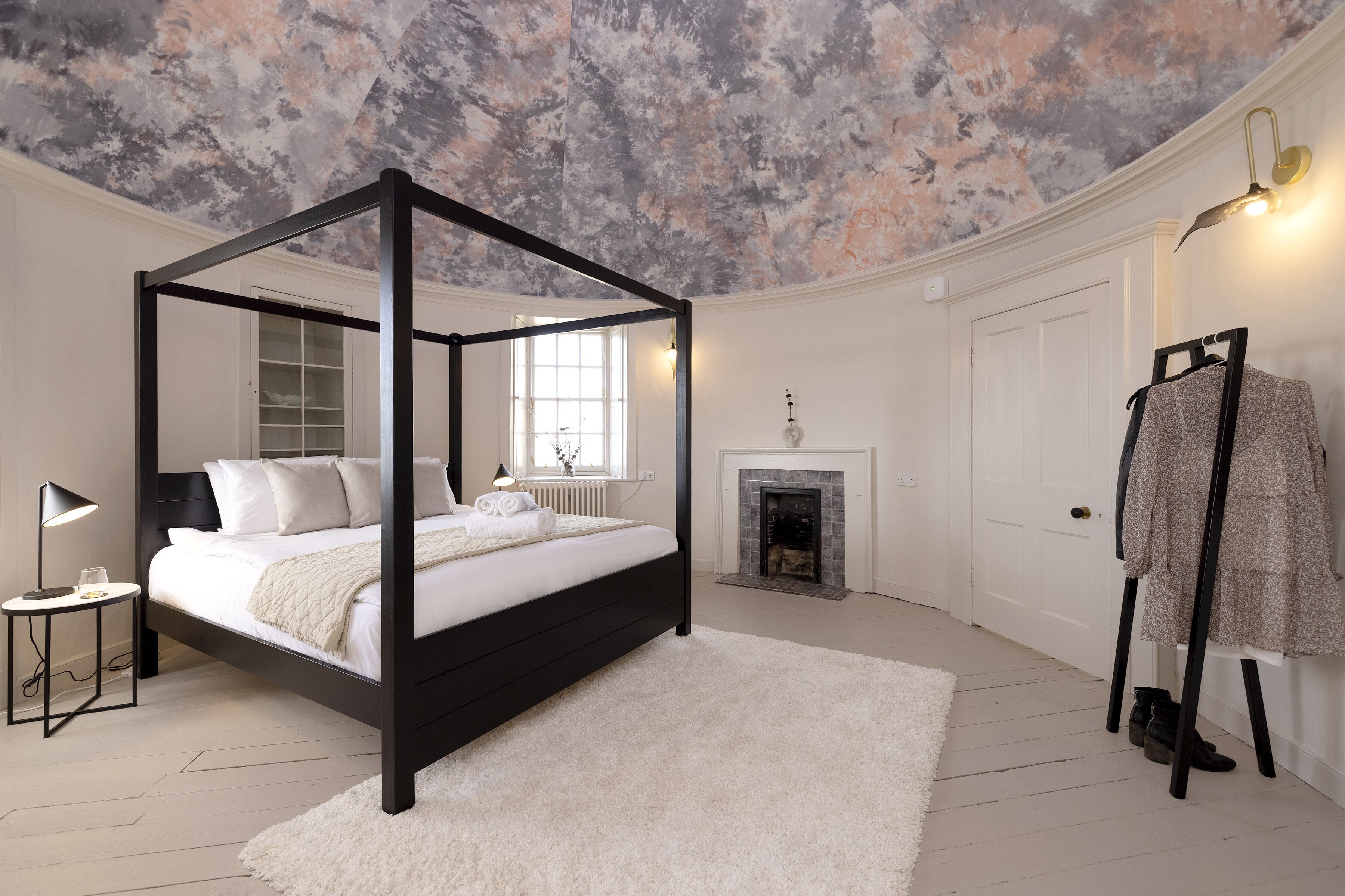 A bedroom with a four-poster bed, a fireplace, a coat rack, and a white shag rug.