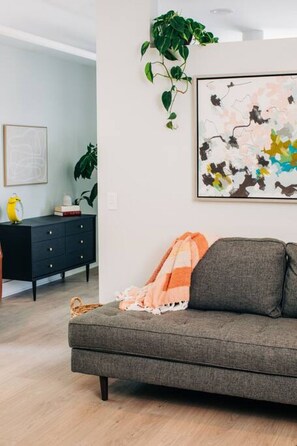 A living room with a grey sofa, a colorful abstract painting, and a potted plant. - Market #10 · Stellar Suites | Queen of Nulu | Boutique Hotel (Louisville)
