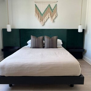 Ein Bett mit weißer Decke, zwei gestreifte Kissen und einem grünen Kopfteil, sowie ein Wandbehang aus Makramee.