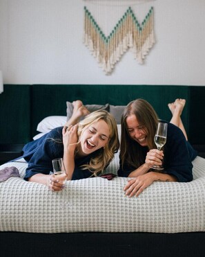 Dos personas reclinadas en una cama con una colcha estampada, una decoración en la pared sobre ellas y una mesita de noche con un vaso.