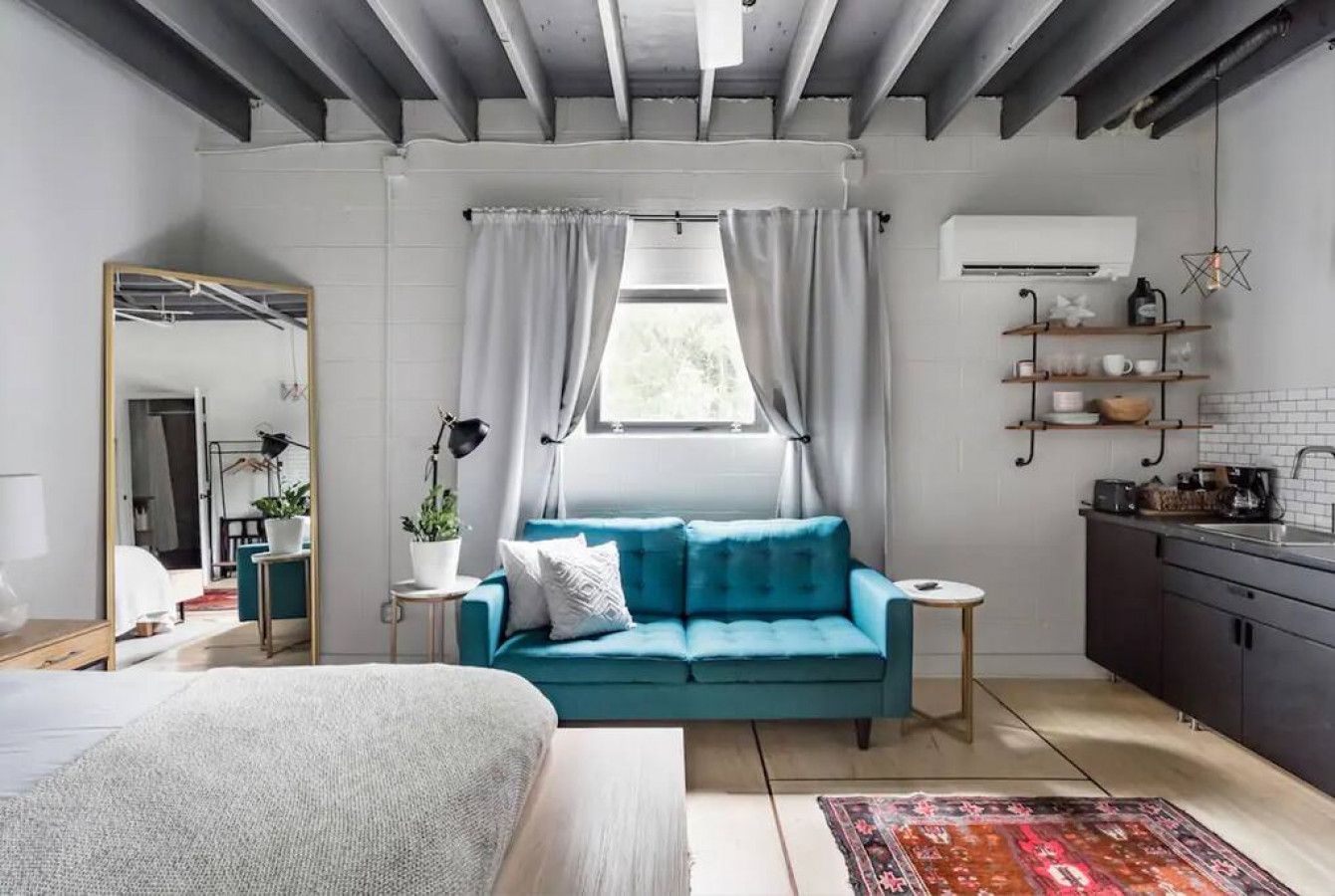 A modern bedroom with a bed, a blue sofa, a small table, a mirror, and a window with curtains.