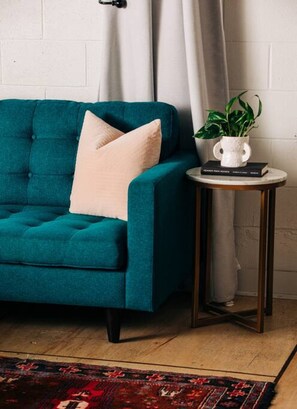 A teal sofa with a beige pillow, a small round table with a white vase and a book, and a patterned rug on a wooden floor.