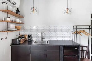 A modern kitchen with a black countertop, white tiled backsplash, and two pendant lights. There are wooden shelves above the counter holding dishes and a coffee maker. - Market #5 · Stellar Suites | Turquoise Suite in NuLu | Bikes (Louisville)
