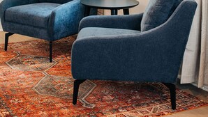 Une pièce avec deux fauteuils bleus, une petite table ronde, un tapis à motifs, une fenêtre avec des rideaux et un tableau abstrait encadré.