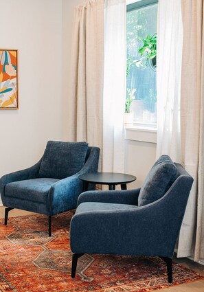 Ein Zimmer mit zwei blauen Sesseln, einem kleinen runden Tisch, einem gemusterten Teppich, einem Fenster mit Vorhängen und einem gerahmten abstrakten Gemälde.