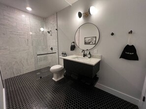 A modern bathroom with a black and white hexagonal tile floor, a white toilet, a dark sink with a round mirror, and a black towel hanging on the wall.