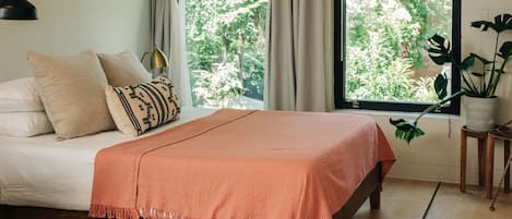 A bedroom with a bed, curtains, a window with a view of greenery, a bedside lamp, and a potted plant.