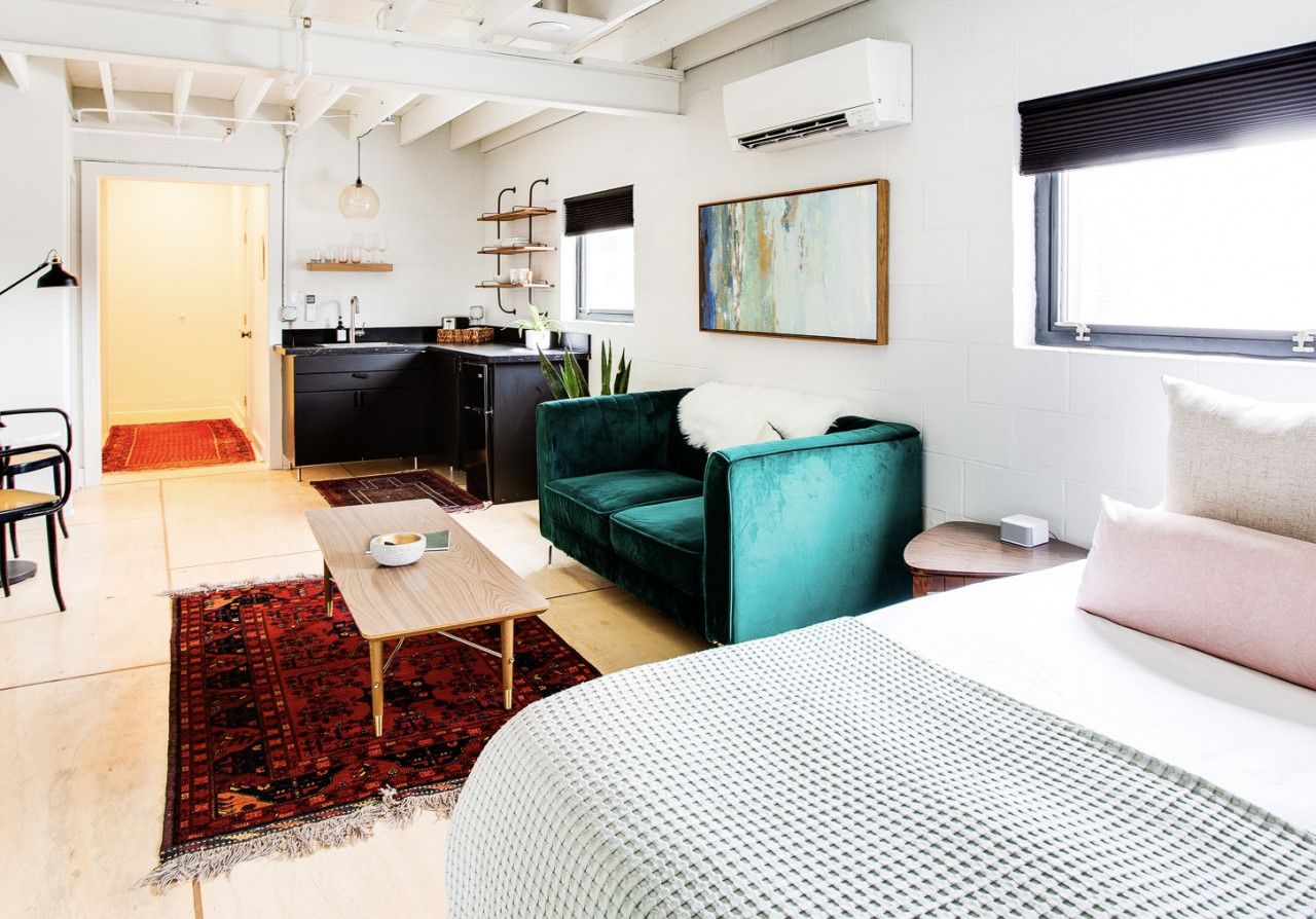 A modern living room with a teal sofa, a wooden coffee table, and a large bed with a white and green patterned bedspread.