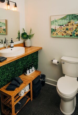 Un baño moderno con tocador de madera, un lavamanos blanco y un inodoro. Hay una pared verde con un mapa de Kentucky, una planta en maceta y una estantería con botellas.