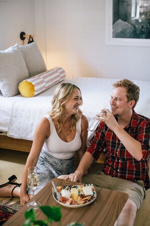 A couple sitting on a bed in a bedroom with a bedside table holding a glass of wine and a plate of food.