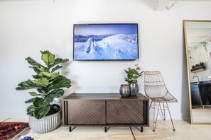 Una sala de estar con un televisor grande en la pared, un mueble de madera, una planta grande en maceta, una silla y un espejo.